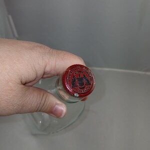 May include: A clear glass bottle with a red metal cap. The cap has a black emblem and text that reads "WALKER & SONS, LTD. HARROW". The bottle is held by a hand.