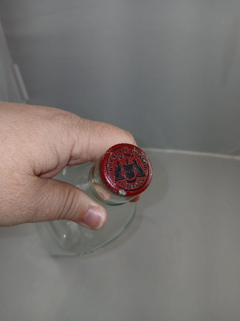 May include: A clear glass bottle with a red metal cap. The cap has a black emblem and text that reads "WALKER & SONS, LTD. HARROW". The bottle is held by a hand.