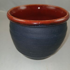 May include: A handmade ceramic bowl with a dark blue exterior and a reddish-brown interior. The rim is also reddish-brown. The bowl has a rounded shape and a textured surface. This pottery bowl is suitable for serving food.
