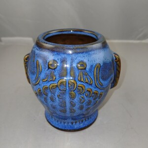 May include: A blue ceramic vase with a round body and a small base. The vase has a textured surface with a pattern of stylized floral or leaf designs in a darker blue and gold color. It has two small handles on the sides.