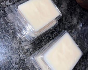 Op de afbeelding: Verschillende stapels vierkante, gebroken witte wassmeltstukken in doorzichtige plastic clamshell verpakkingen. De wassmeltstukken hebben een glad oppervlak en zijn gerangschikt op een donker, gespikkeld aanrechtblad. Een houten bak is zichtbaar op de achtergrond.