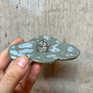 May include: A ceramic cloud-shaped ring dish with a speckled blue-gray glaze. A silver ring with a light-colored stone sits on the dish. The dish is held in a hand.