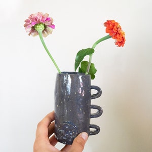 Pode incluir: Um vaso de cerâmica cinza escuro com três alças de laço e um acabamento salpicado. O vaso contém duas zínias coloridas, uma rosa e outra laranja, com caules e folhas verdes. O vaso é segurado contra um fundo branco.