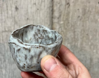 Op de afbeelding: Een kleine, handgemaakte keramische kom met een getextureerd, gespikkeld oppervlak. De kom is voornamelijk wit met bruine en grijze accenten. De rand heeft een onregelmatige, organische vorm. De kom wordt in een hand gehouden.