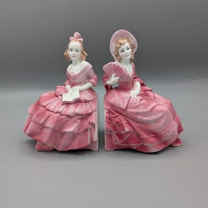 May include: Two porcelain figurines of women in pink dresses. One holds a book, the other a fan. The figures are seated, with detailed dresses and hairstyles. The figurines are likely decorative items.
