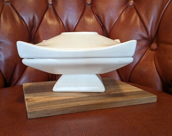 May include: A white ceramic serving dish with a lid, featuring a unique, modern design. The dish is sitting on a wooden base.