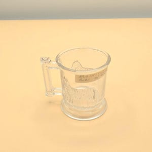 May include: Clear glass beer mug with a handle and etched design of a horse. The mug has the words "Another Baby" written on a label. The mug is sitting on a light tan surface.