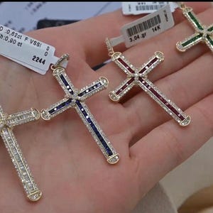Puede incluir: Tres colgantes de cruz de oro con piedras preciosas de diferentes colores. La cruz de la izquierda tiene piedras azules, la cruz del medio tiene piedras rojas y la cruz de la derecha tiene piedras verdes. Cada cruz est&aacute; engastada con piedras transparentes m&aacute;s peque&ntilde;as.