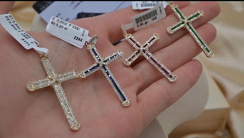 Puede incluir: Tres colgantes de cruz de oro con piedras preciosas de diferentes colores. La cruz de la izquierda tiene piedras azules, la cruz del medio tiene piedras rojas y la cruz de la derecha tiene piedras verdes. Cada cruz est&aacute; engastada con piedras transparentes m&aacute;s peque&ntilde;as.