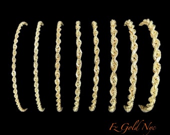 May include: A set of seven gold rope chains of varying thicknesses, displayed against a black background.  The chains are arranged in a row, with the thickest chain on the left and the thinnest chain on the right.  The chains are all made of a shiny, polished gold.