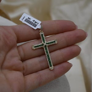 Puede incluir: Un colgante de cruz de oro con piedras preciosas verdes engastadas en el centro y a lo largo de los brazos. La cruz est&aacute; sobre un fondo blanco.