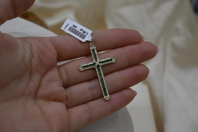 Puede incluir: Un colgante de cruz de oro con piedras preciosas verdes engastadas en el centro y a lo largo de los brazos. La cruz est&aacute; sobre un fondo blanco.