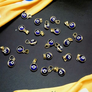 Puede incluir: Una colecci&oacute;n de 20 colgantes de oro de ojo turco con esmalte azul y blanco. Los colgantes est&aacute;n dispuestos sobre una superficie negra con una tela amarilla en el fondo.