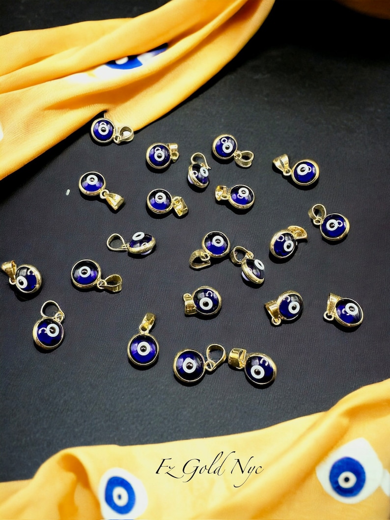 Puede incluir: Una colecci&oacute;n de 20 colgantes de oro de ojo turco con esmalte azul y blanco. Los colgantes est&aacute;n dispuestos sobre una superficie negra con una tela amarilla en el fondo.