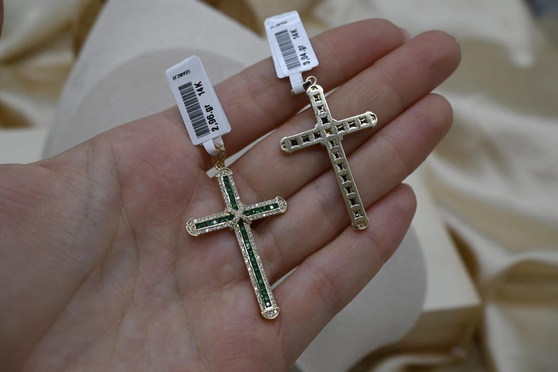 Puede incluir: Dos colgantes de cruz de oro con piedras preciosas verdes. La cruz de la izquierda es m&aacute;s peque&ntilde;a y est&aacute; adornada con piedras preciosas m&aacute;s peque&ntilde;as. La cruz de la derecha es m&aacute;s grande y tiene piedras preciosas m&aacute;s grandes.