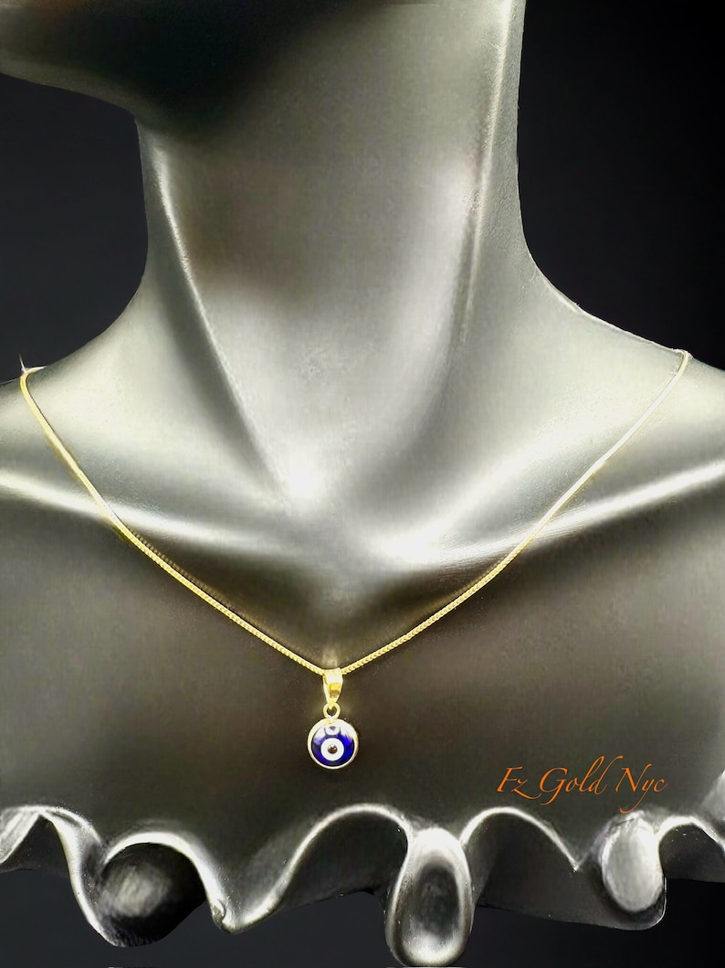 Puede incluir: Un collar de cadena de oro con un colgante azul de ojo turco. El colgante es redondo y tiene un centro blanco. El collar est&aacute; en un maniqu&iacute; negro.