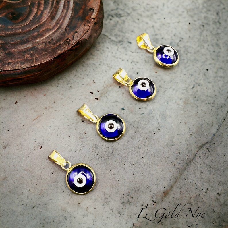 Puede incluir: Cuatro colgantes de oro con ojo turco de vidrio azul y blanco. Los colgantes est&aacute;n sobre una superficie gris texturizada.