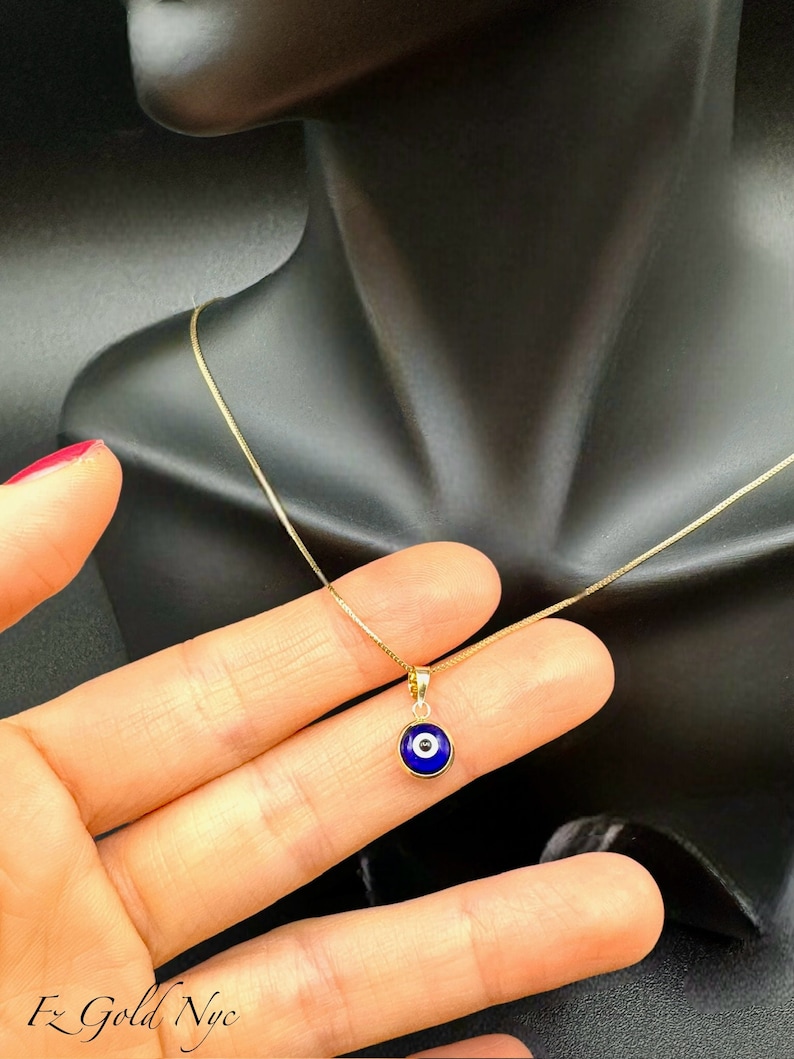 Puede incluir: Un collar de cadena de oro con un colgante de ojo turco azul. El colgante es redondo y tiene un centro blanco.