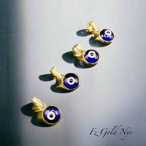 Puede incluir: Cuatro colgantes de oro con ojo turco de vidrio azul y blanco. Los colgantes est&aacute;n dispuestos en fila sobre un fondo gris.