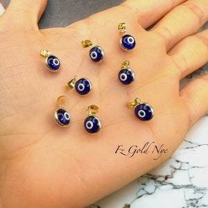 Puede incluir: Ocho colgantes de oro con un mal de ojo de esmalte azul y blanco. Los colgantes est&aacute;n en una mano.
