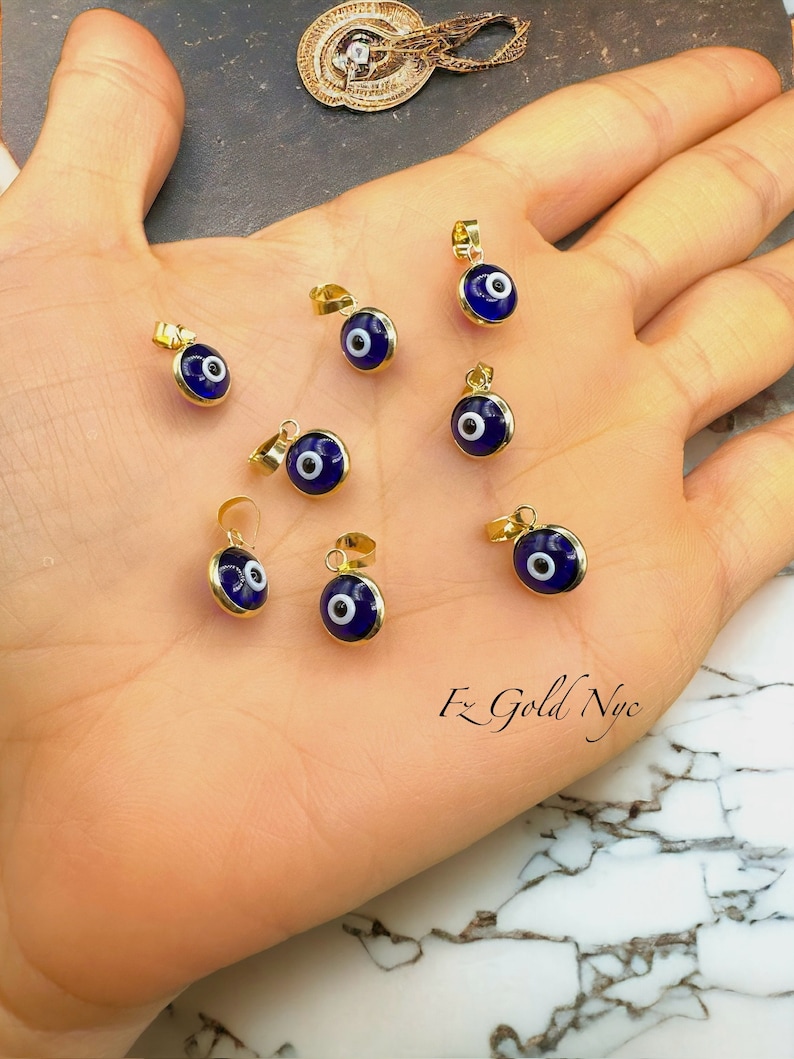 Puede incluir: Ocho colgantes de oro con un mal de ojo de esmalte azul y blanco. Los colgantes est&aacute;n en una mano.
