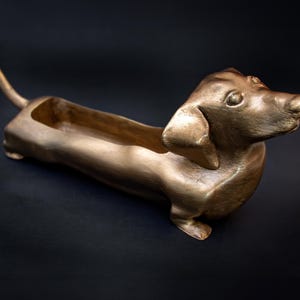 May include: A gold-colored dachshund planter with a long, narrow body and a raised head. The planter has a hollow interior for planting.