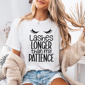 May include: White t-shirt with the text "Lashes longer than my patience" in black. The shirt features a graphic of eyelashes above the text. The model is wearing denim shorts and a beige cardigan.