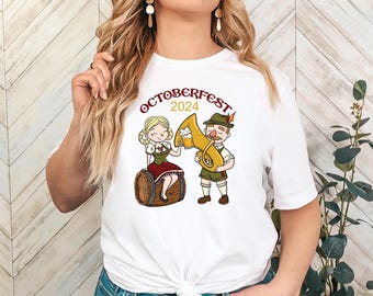 May include: White t-shirt featuring a cartoon illustration for Oktoberfest 2024. The design includes a woman in a traditional dress sitting on a barrel and a man with a horn and beer. The text "OCTOBERFEST 2024" is arched above the figures.