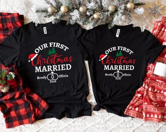 Può includere: Due magliette nere a maniche corte con la scritta bianca e rossa "OUR FIRST Christmas MARRIED", un cappello da Babbo Natale, alberi verdi e due anelli intrecciati con i nomi "Brody" e "Olivia" e l'anno "2026" stampati in bianco.