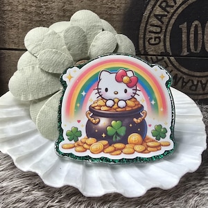 Pode incluir: Um autocolante colorido com Hello Kitty sentada num pote de ouro no final de um arco-íris. O autocolante tem uma borda verde brilhante e trevos. O fundo é uma superfície branca e enrugada.