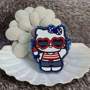 May include: A glittery blue and white Hello Kitty pin. The pin features Hello Kitty wearing heart-shaped sunglasses, a red, white, and blue striped shirt, and blue shorts. She is holding an ice pop. The pin is on a white seashell.