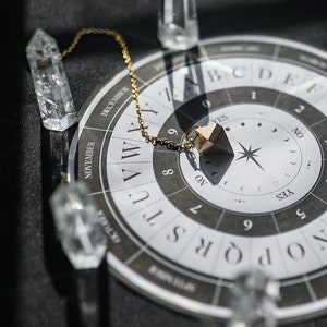 May include: A gold pendulum with a chain hangs over a black and white circular chart with letters, numbers, and the words "YES", "NO", and "ON".  The chart is surrounded by six clear quartz crystals.