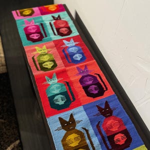 May include: A colorful, rectangular woven table runner featuring a repeating pattern of stylized cat figures. Each cat is depicted in a square, with a variety of vibrant colors including pink, green, blue, and red. The runner is placed on a dark brown surface.