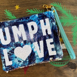 May include: A sequined clutch with a blue tie-dye pattern and the words "UMPH LOVE" in white. The clutch has a light blue wrist strap and a gold-colored zipper pull. The background is a wooden surface with painted tropical designs.