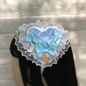 Może przedstawiać: Jasnoniebieska opaska do włos&oacute;w w kształcie serca z białą koronką i dwoma niebieskimi kokardkami z satyny. Opaska ma mały złoty zawieszkę w środku.