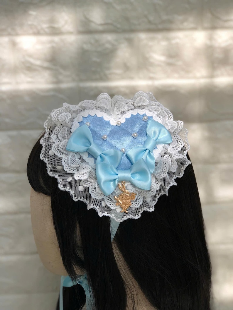 Może przedstawiać: Jasnoniebieska opaska do włos&oacute;w w kształcie serca z białą koronką i dwoma niebieskimi kokardkami z satyny. Opaska ma mały złoty zawieszkę w środku.