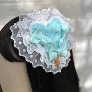 Może przedstawiać: Spinka do włos&oacute;w w kształcie serca w jasnoniebieskim kolorze, ozdobiona białą koronką i perłowymi akcentami. Spinka ma dwie jasnoniebieskie satynowe kokardki i złoty zawieszkę.