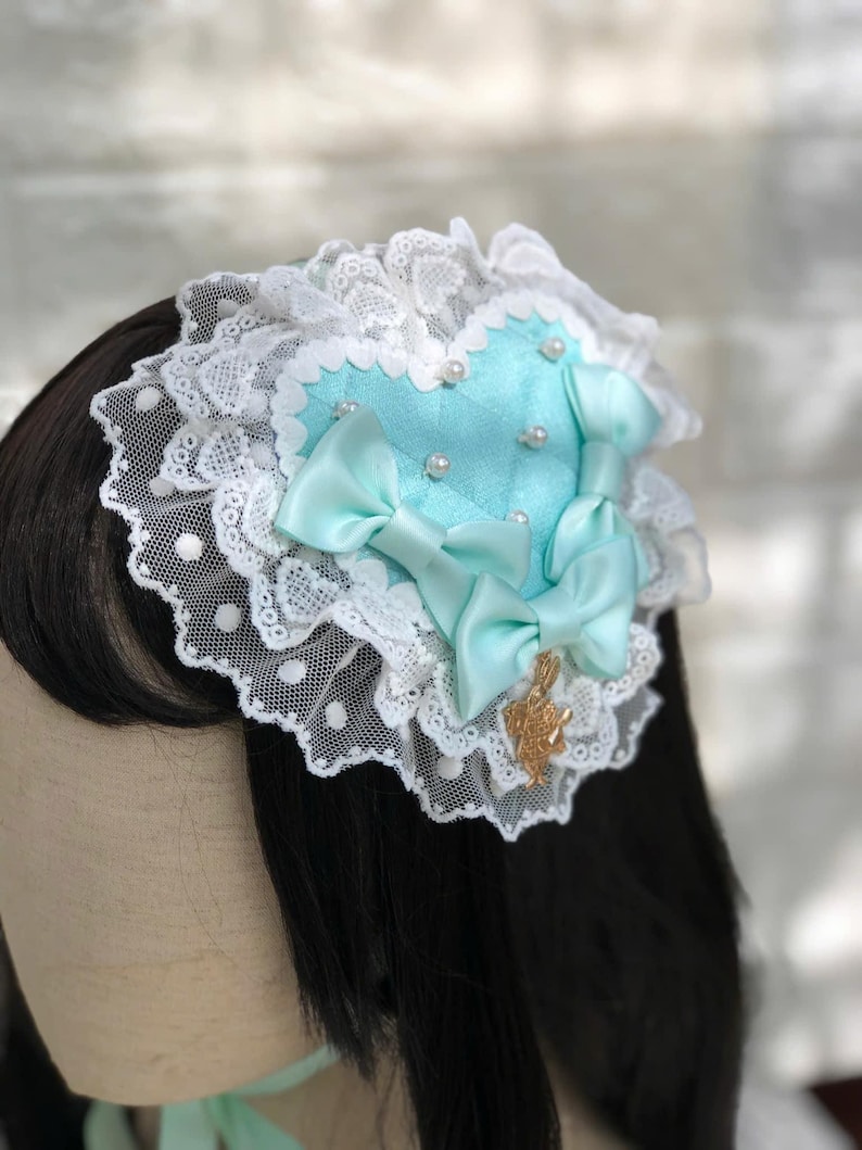 Może przedstawiać: Spinka do włos&oacute;w w kształcie serca w jasnoniebieskim kolorze, ozdobiona białą koronką i perłowymi akcentami. Spinka ma dwie jasnoniebieskie satynowe kokardki i złoty zawieszkę.