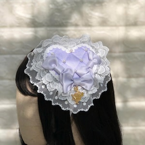 Może przedstawiać: Fioletowa spinka do włos&oacute;w w kształcie serca z białą koronką i złotą kokardką. Spinka ma perłową ozdobę i mały złoty zawieszkę.