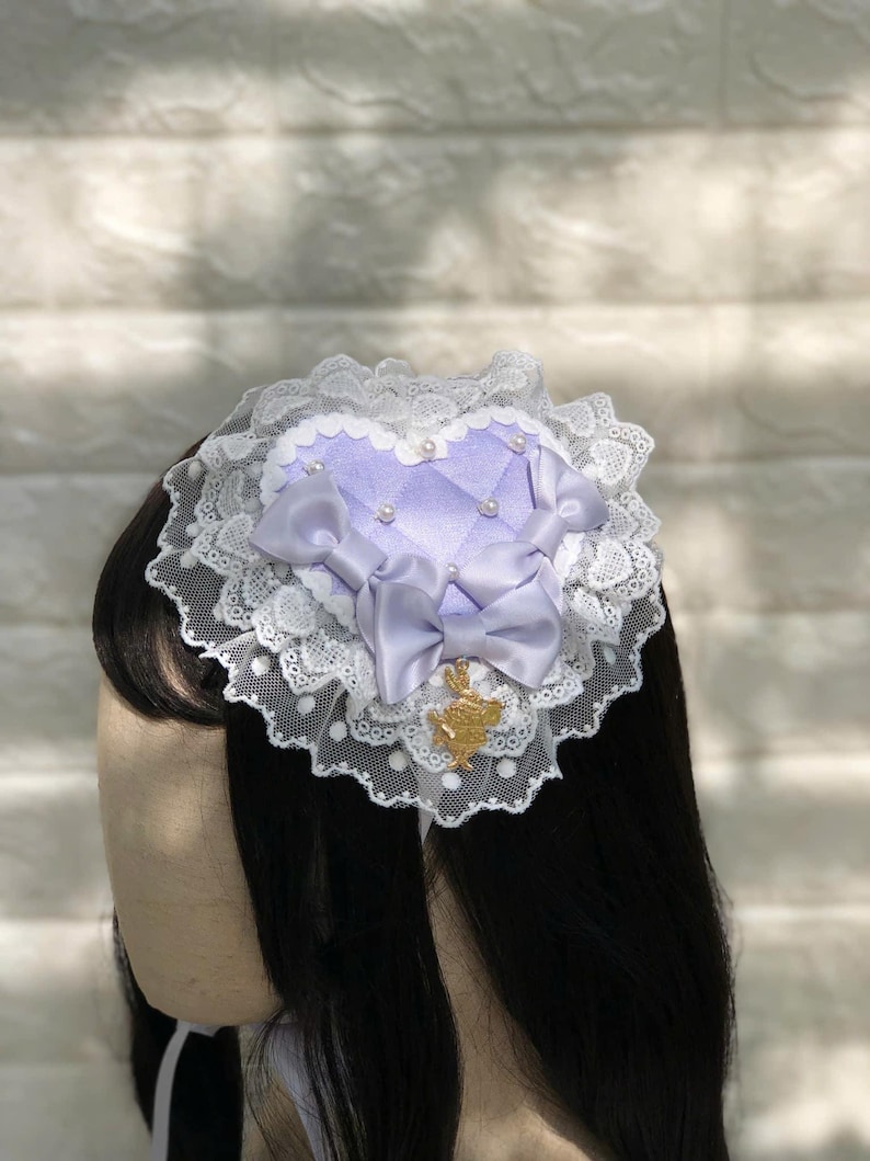 Może przedstawiać: Fioletowa spinka do włos&oacute;w w kształcie serca z białą koronką i złotą kokardką. Spinka ma perłową ozdobę i mały złoty zawieszkę.