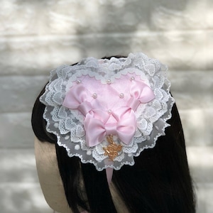 Może przedstawiać: R&oacute;żowa opaska do włos&oacute;w w kształcie serca z białą koronką i złotym akcentem. Opaska ozdobiona jest r&oacute;żowymi satynowymi kokardkami i perłami.