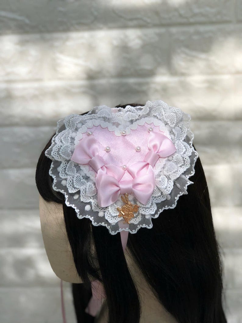 Może przedstawiać: R&oacute;żowa opaska do włos&oacute;w w kształcie serca z białą koronką i złotym akcentem. Opaska ozdobiona jest r&oacute;żowymi satynowymi kokardkami i perłami.