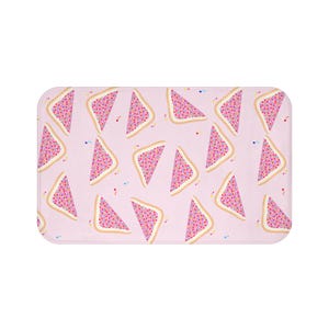 Puede incluir: Una alfombra de baño rectangular con un patrón repetido de triángulos rosas que se asemejan a rebanadas de pan tostado con chispas de colores sobre un fondo rosa claro. La alfombra tiene esquinas redondeadas.