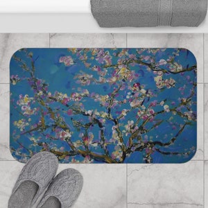 May include: Rectangular bath mat featuring a vibrant floral painting in shades of blue, pink, and white. The artwork depicts a blossoming tree against a bright blue background. The mat is placed on a tiled floor, with a pair of gray slippers visible.