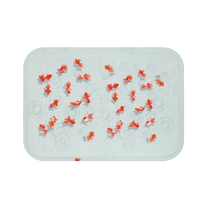 Puede incluir: Una alfombra de baño azul claro con un patrón de peces rojos y círculos blancos. La alfombra es rectangular y tiene una superficie suave y texturizada.