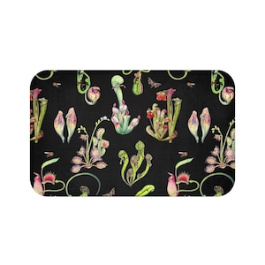 May include: Rectangular black bath mat featuring a repeating pattern of watercolor carnivorous plants, including pitcher plants and Venus flytraps. The plants are in shades of green, pink, and red, with small insects scattered throughout the design.