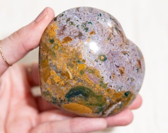 May include: A heart-shaped polished stone with a variety of colors, including purple, white, yellow, and green. The stone has a smooth, polished surface and is a natural, organic material.