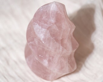 May include: A pink rose quartz crystal carved into a spiral shape. The crystal is smooth and polished, with a light pink hue.