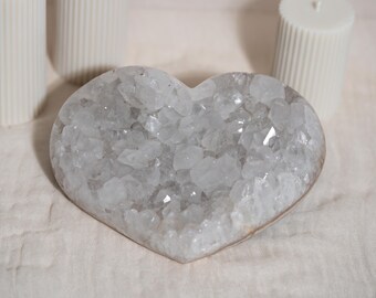 May include: A white heart-shaped crystal with a cluster of smaller crystals inside. The crystal is on a white fabric surface.