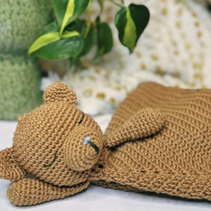 May include: A crocheted brown teddy bear with closed eyes is lying on a brown crocheted blanket. The blanket is folded over the bear's body.